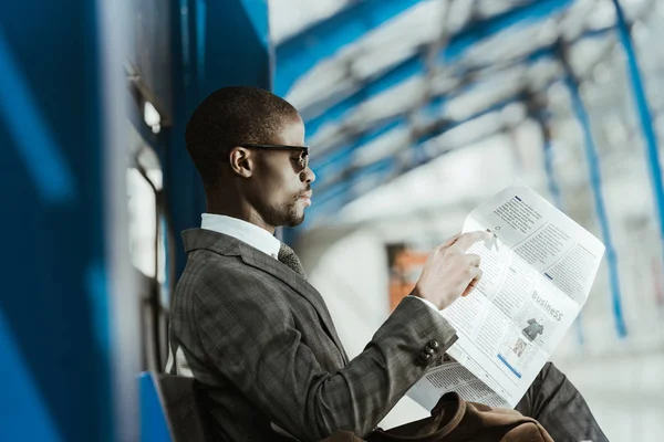 Homme d'affaires afro-américain portant costume lecture journal sur banc — Photo de stock