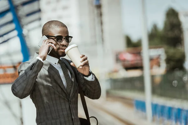 Hombre de negocios afroamericano que usa traje con teléfono inteligente y taza de café esperando el tren - foto de stock