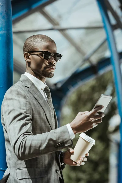 Stylish confident businessman with smartphone and coffee cup waiting for train — Stock Photo