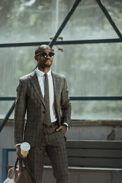 Stylish confident businessman with briefcase and coffee cup standing on public transport station — Stock Photo