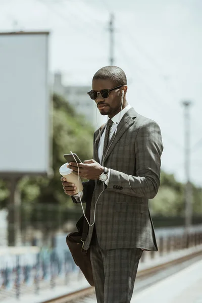 Junger afrikanisch-amerikanischer Geschäftsmann hört Musik und hält Kaffeetasse auf dem Bahnhof — Stockfoto