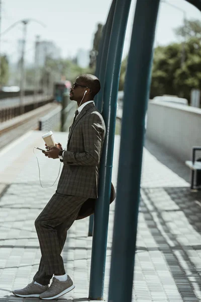 Stylish confident businessman listening to music and holding coffee cup on train station — Stock Photo