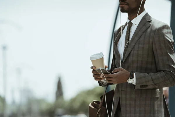 Vista recortada del hombre de negocios afroamericano que usa traje escuchando música y sosteniendo la taza de café en la estación de tren - foto de stock