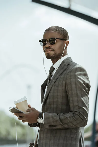 Young african american businessman listening to music and holding coffee cup on public transport station — Stock Photo