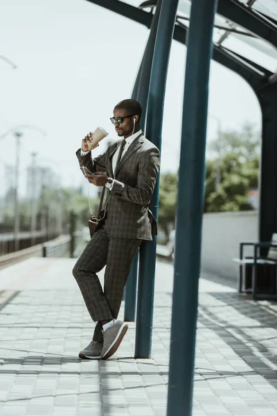 Sonriente empresario confiado escuchando música y sosteniendo la taza de café en la estación de transporte público - foto de stock