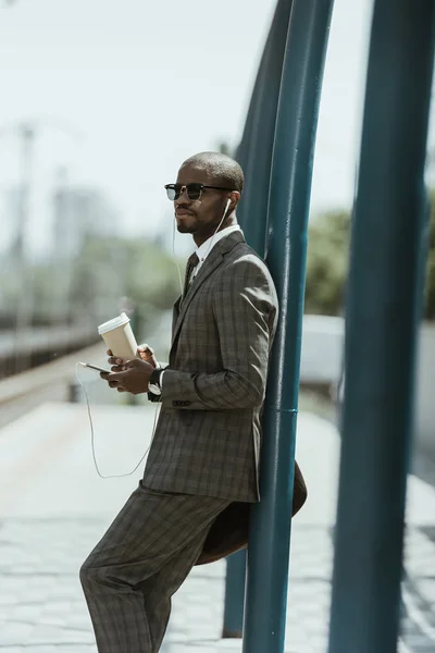 Empresario afroamericano con traje escuchando música y sosteniendo taza de café en la estación de transporte público - foto de stock