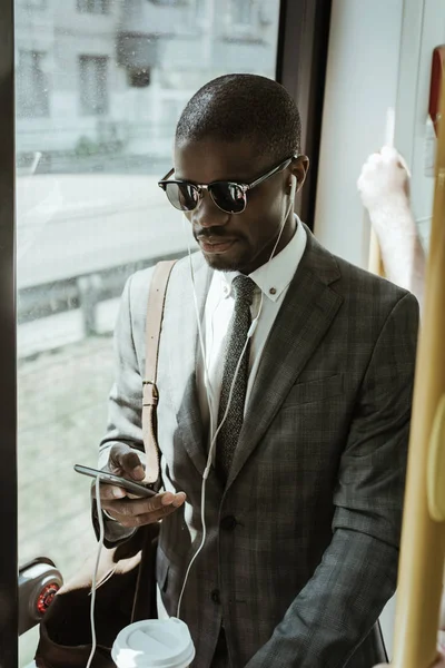 African american businessman wearing suit in earphones using smartphone while taking train — Stock Photo
