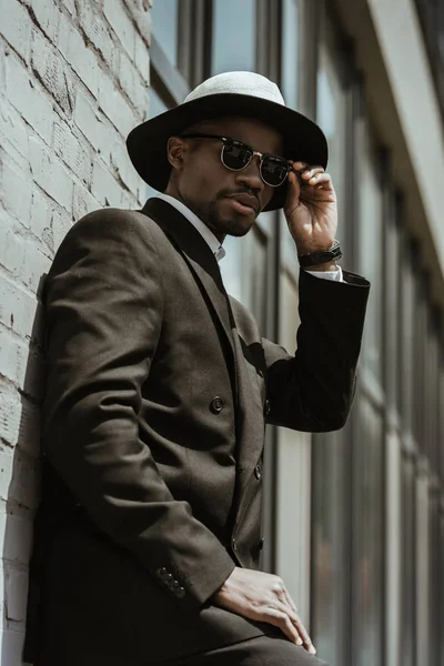 Modischer afrikanisch-amerikanischer Geschäftsmann mit Fedora-Hut und Sonnenbrille posiert an einer Hauswand — Stockfoto
