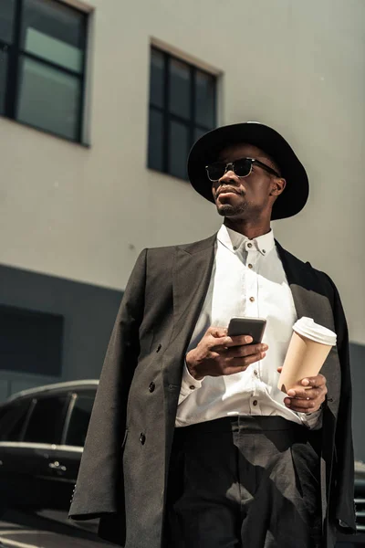 Moderno hombre de negocios afroamericano que usa sombrero de sombrero de sombrero de sombrero de fedora con teléfono inteligente y taza de café - foto de stock