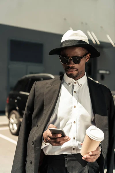 Bello giovane uomo africano americano utilizzando smartphone e tenendo in mano la tazza di caffè — Foto stock