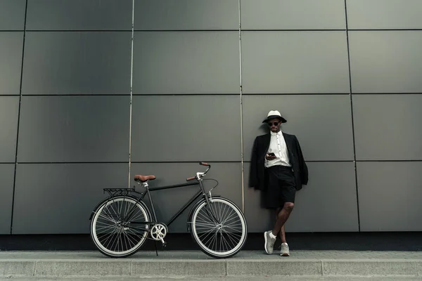 El hombre afroamericano de moda que usa sombrero de sombrero de fedora usando un teléfono inteligente mientras está de pie junto a su bicicleta - foto de stock