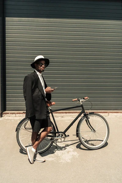 Elegante hombre con camisa blanca y chaqueta usando smartphone mientras sostiene su bicicleta - foto de stock