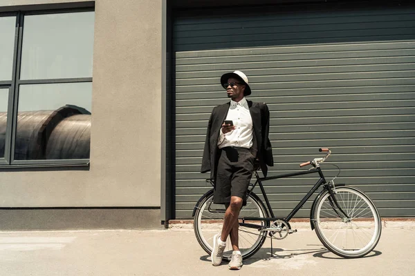 Стильний чоловік у білій сорочці та піджаку використовує смартфон, спираючись на свій велосипед — стокове фото