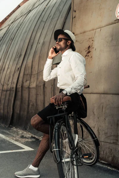 Stylish man wearing white shirt leaning on bike and talking on smartphone — Stock Photo