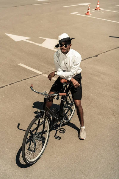 Fashionable african american man wearing fedora hat and sunglasses posing by city bike — Stock Photo