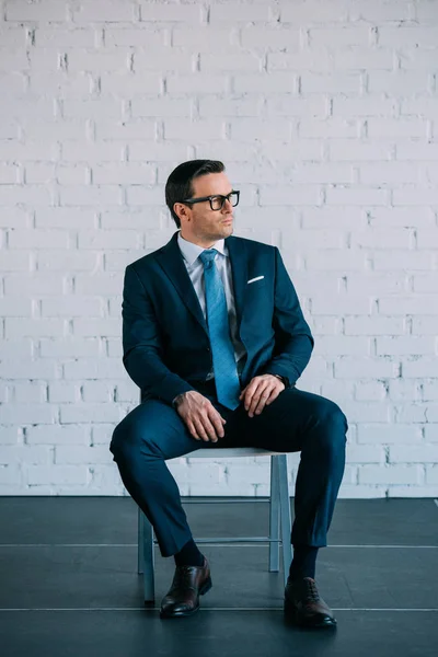 Serious middle aged businessman in suit and eyeglasses sitting on chair and looking away — Stock Photo