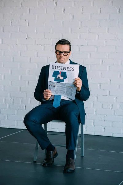 Серьезный бизнесмен средних лет в формальной одежде и очках сидит на стуле и читает газету — стоковое фото