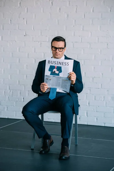 Serio hombre de negocios de mediana edad en ropa formal y anteojos sentados en la silla y leyendo el periódico - foto de stock