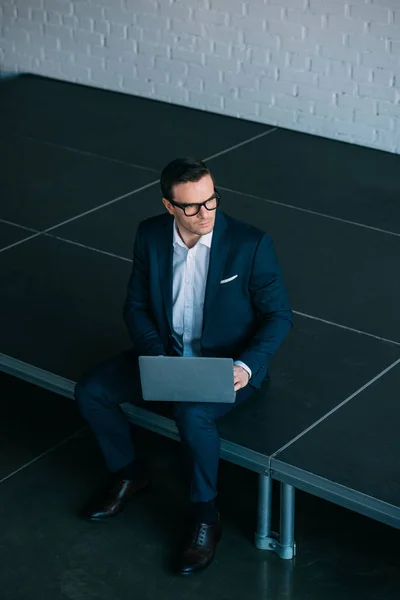 Vista de ángulo alto de hombre de negocios de mediana edad en traje y gafas usando el ordenador portátil y mirando hacia otro lado - foto de stock