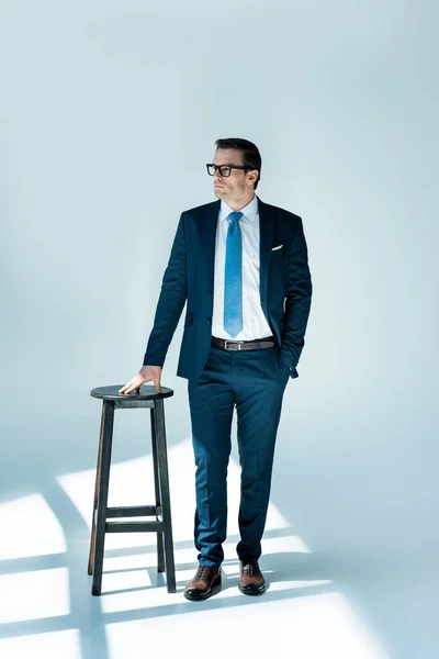 Full length view of serious buisnessman in eyeglasses standing near stool and looking away on white — Stock Photo