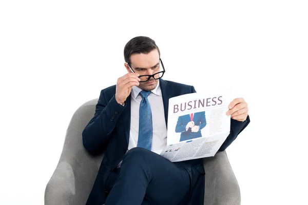 Зосереджений бізнесмен регулює окуляри і читає газету, сидячи в кріслі ізольовано на білому — стокове фото