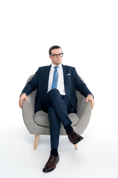 Full length view of serious middle aged businessman in eyeglasses and formal wear sitting in armchair and looking away on white — Stock Photo