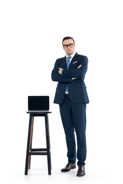 Confiado hombre de negocios de pie con los brazos cruzados cerca de la computadora portátil en el taburete blanco - foto de stock
