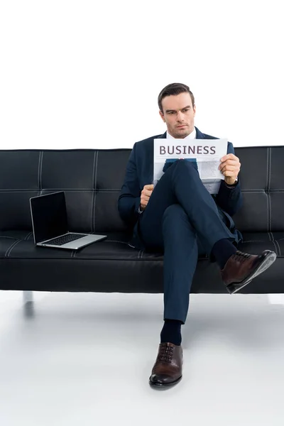 Vista completa del empresario de mediana edad leyendo el periódico mientras está sentado en el sofá con el ordenador portátil en blanco - foto de stock