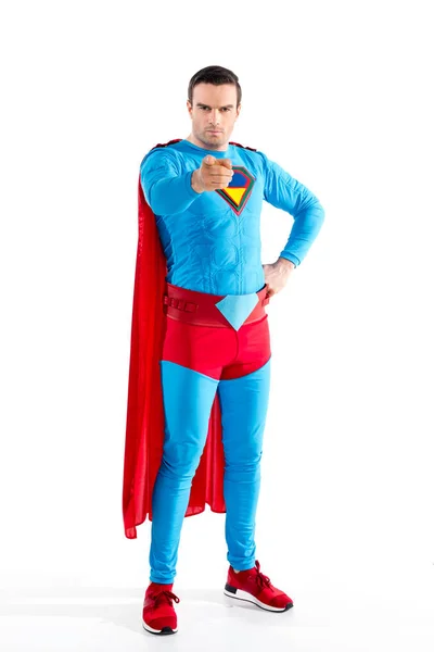 Vista de longitud completa de superhéroe masculino seguro de pie con la mano en la cintura y apuntando a la cámara aislada en blanco - foto de stock