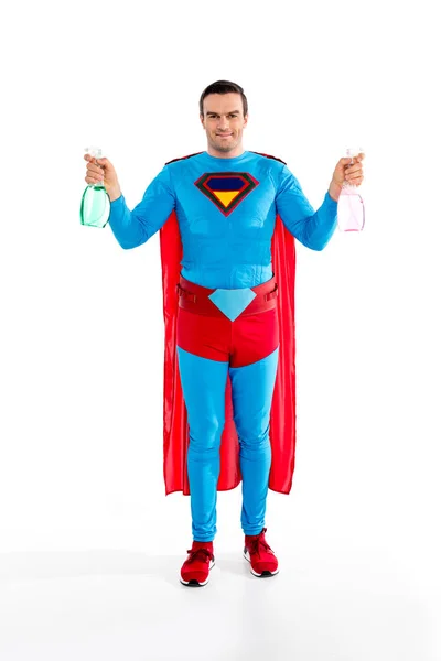 Guapo superman sosteniendo botellas de spray y sonriendo a la cámara aislado en blanco - foto de stock