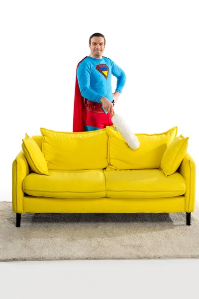 Handsome man in superhero costume cleaning couch with duster and smiling at camera on white — Stock Photo