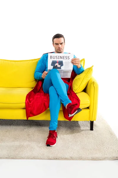 Полный вид серьезного мужского супергероя, сидящего на диване и читающего деловую газету в белом — стоковое фото