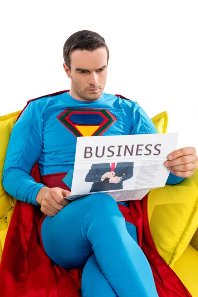 Sério super-herói masculino sentado no sofá e lendo jornal de negócios isolado no branco — Fotografia de Stock