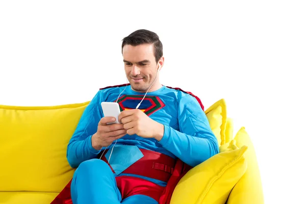 Smiling superman in earphones sitting on sofa and using smartphone isolated on white — Stock Photo