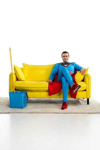 Superhéroe masculino sonriente sentado en el sofá cerca de cubo y fregona en blanco - foto de stock