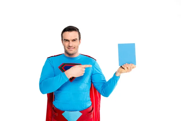 Guapo superhéroe masculino apuntando con el dedo a la caja en blanco con detergente y sonriendo a la cámara aislada en blanco - foto de stock