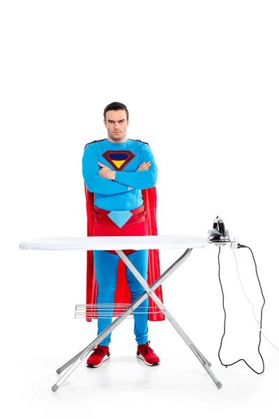 Handsome superman standing with crossed arms near ironing board and looking at camera on white — Stock Photo