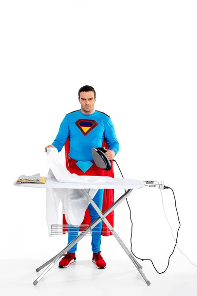 Man in superhero costume ironing clothes and looking at camera on white — Stock Photo