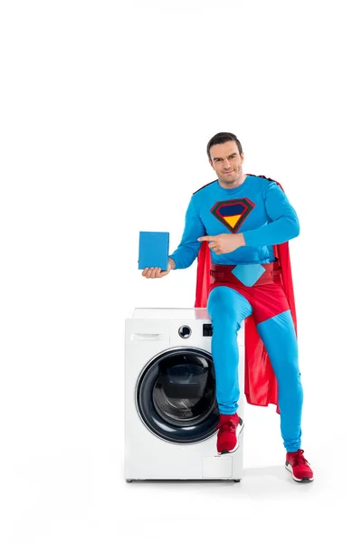 Smiling male superhero sitting on washing machine and pointing with finger at soap powder on white — Stock Photo