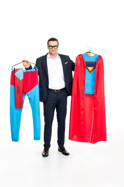 Businessman in eyeglasses holding hangers with superhero costume and smiling at camera isolated on white — Stock Photo