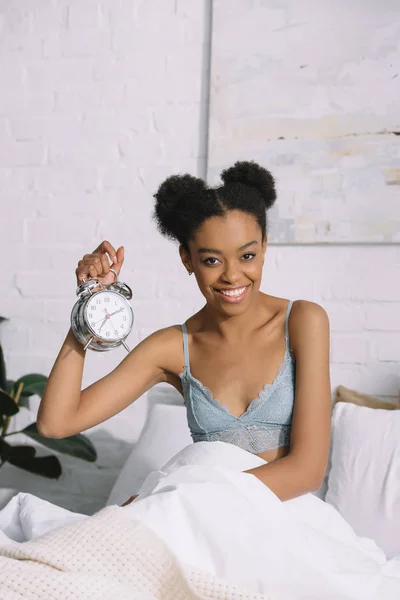 Chica afroamericana sonriente sosteniendo el despertador mientras está sentada en la cama por la mañana - foto de stock