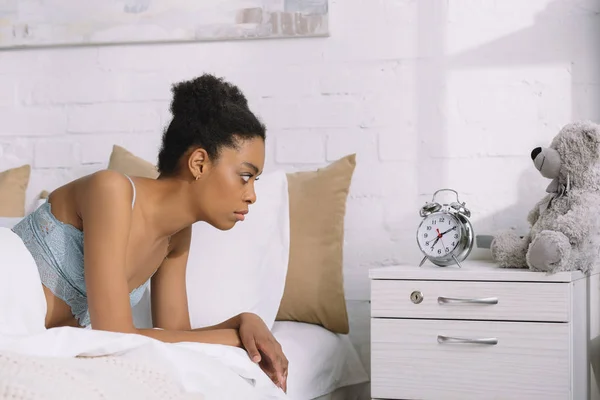 Mujer afroamericana molesta mirando el despertador mientras está acostada en la cama - foto de stock