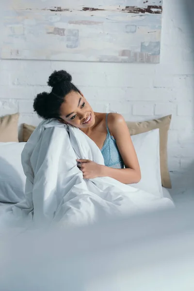 Joven sonriente afroamericana mujer sentada en la cama en la mañana - foto de stock