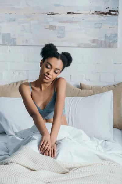 Tierna afroamericana americana chica con los ojos cerrados sentado en la cama en la mañana - foto de stock