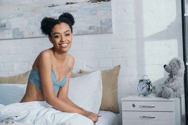 Улыбающаяся африканская американская девушка, сидящая на кровати утром — стоковое фото