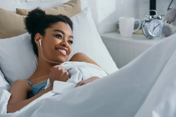Heureux afro-américaine fille écouter de la musique avec des écouteurs tout en étant couché dans le lit — Photo de stock