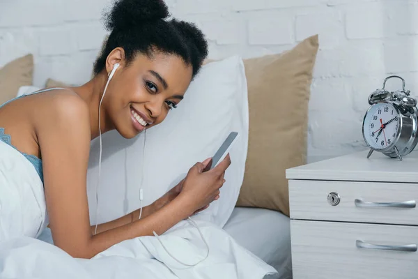 Sorridente ragazza afroamericana ascoltare musica con auricolari e utilizzando smartphone, sveglia sul comodino vicino — Foto stock
