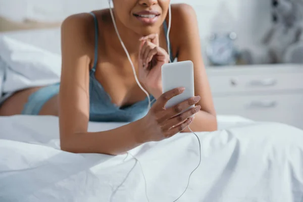 Vue recadrée de fille afro-américaine écoutant de la musique avec des écouteurs et utilisant un smartphone sur le lit — Photo de stock