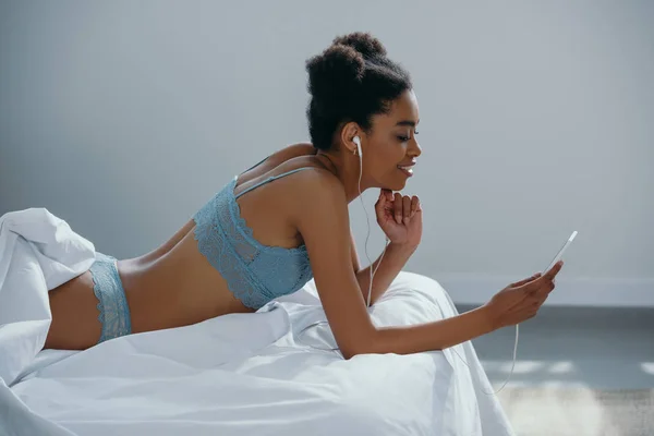 Belle fille afro-américaine en lingerie écouter de la musique avec des écouteurs et en utilisant un smartphone sur le lit le matin — Photo de stock