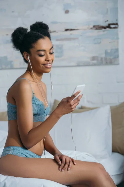 Sorrindo menina americana africana em lingerie ouvindo música com fones de ouvido e smartphone de manhã — Fotografia de Stock
